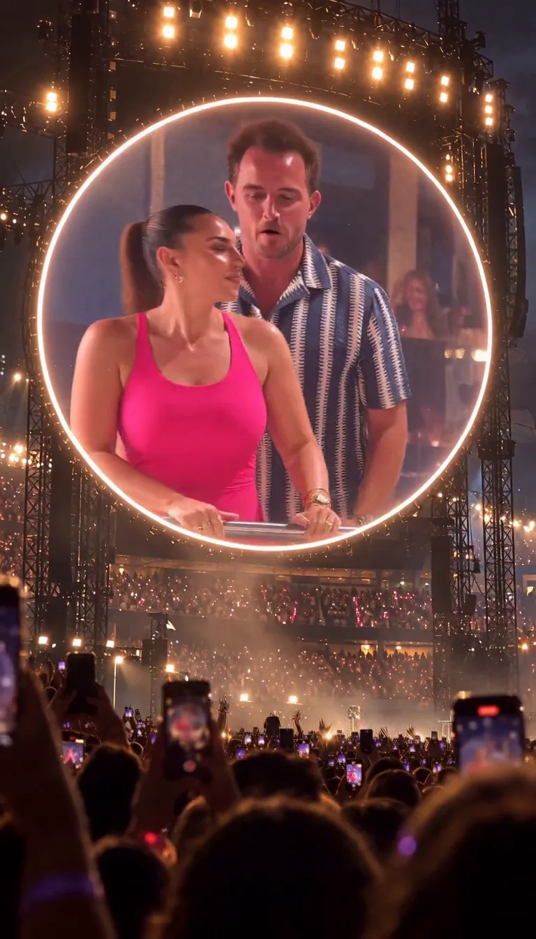 On a round stadium screen a man in a striped shirt and a woman in pink stand at the railing the moment they realise they are on the kiss cam.
